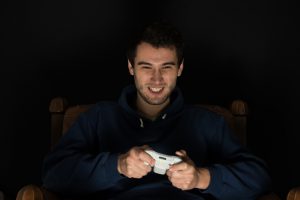 Young man plays computer games sitting in dark room in front of tv or monitor, holding game controller in his hands